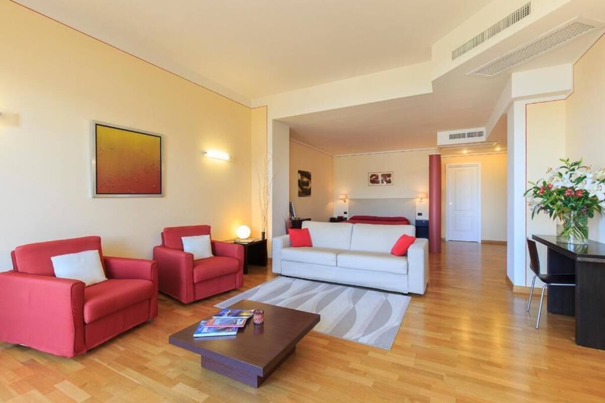 A modern living room with two red sofas, a white sofa, and a wooden coffee table. There is a painting on the wall and a plant on the right side.