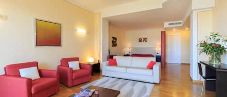 A modern living room with two red sofas, a white sofa, and a wooden coffee table. There is a painting on the wall and a plant on the right side.