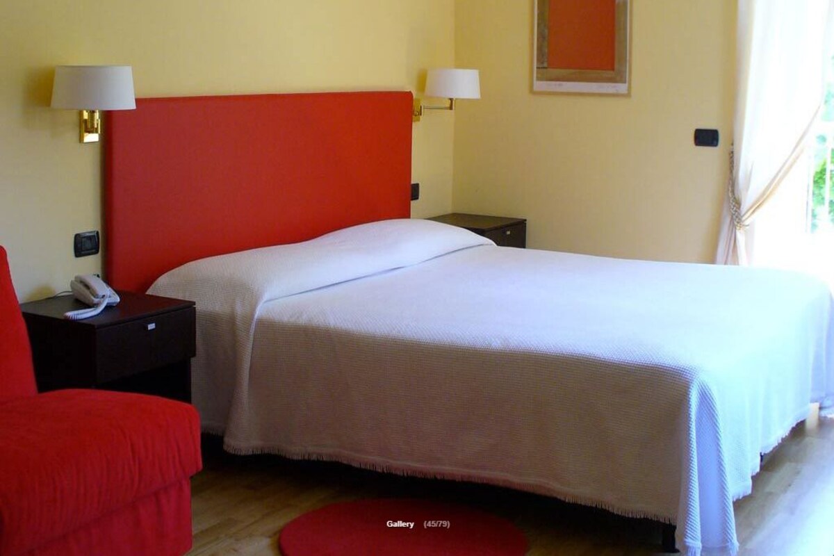 Habitación de hotel con cabecera roja, ropa de cama blanca, una silla roja y una mesita de noche pequeña con una lámpara.