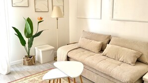 A modern living room with a beige sofa, a white coffee table, and a floor lamp. There are abstract wall art pieces and a large plant with green leaves. - Chez Grand-Mère unique experience, close to the center, free parking (Metz)