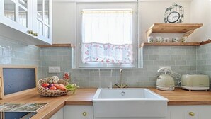 A kitchen with a white sink, wooden countertop, and a window with a floral curtain. There is a basket of vegetables on the counter and a clock on the shelf above. - Chez Grand-Mère unique experience, close to the center, free parking (Metz)