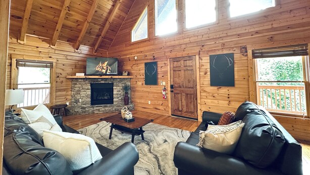 A cozy cabin interior with a stone fireplace, wooden walls, a black leather sofa, and a large window.