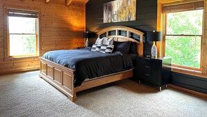 A wooden cabin bedroom with a bed, two bedside tables with lamps, a large window, and a ceiling with exposed beams.