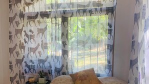 A room with a bed, patterned curtains, a window with a view of greenery, and a small table with a plant.