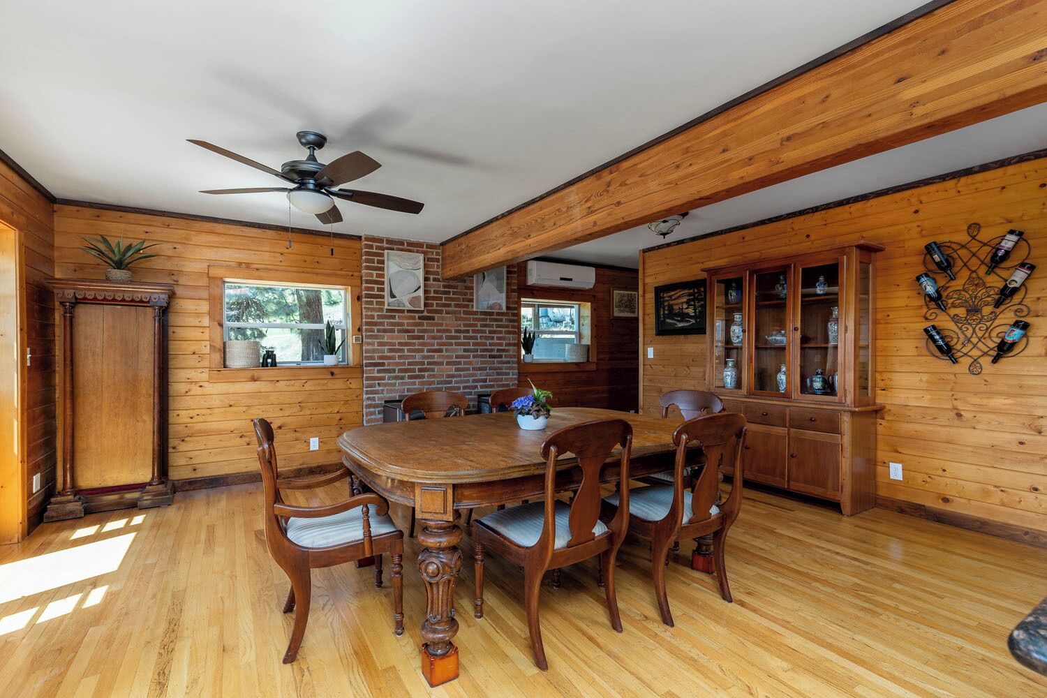 A wooden interior with a dining table, chairs, a ceiling fan, a brick wall, and a cabinet with bottles.