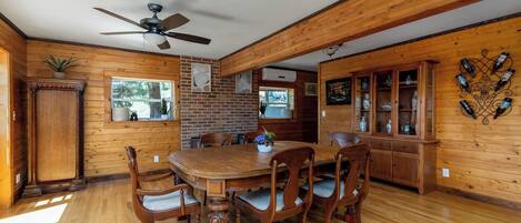A wooden interior with a dining table, chairs, a ceiling fan, a brick wall, and a cabinet with bottles.