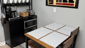Une cuisine compacte équipée d’une machine à café, d’un grille-pain et d’un four à micro-ondes. On y trouve une table recouverte d’une nappe à rayures et une chaise.