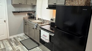 Une cuisine de taille réduite avec des meubles gris, un réfrigérateur noir et une fenêtre équipée de stores.