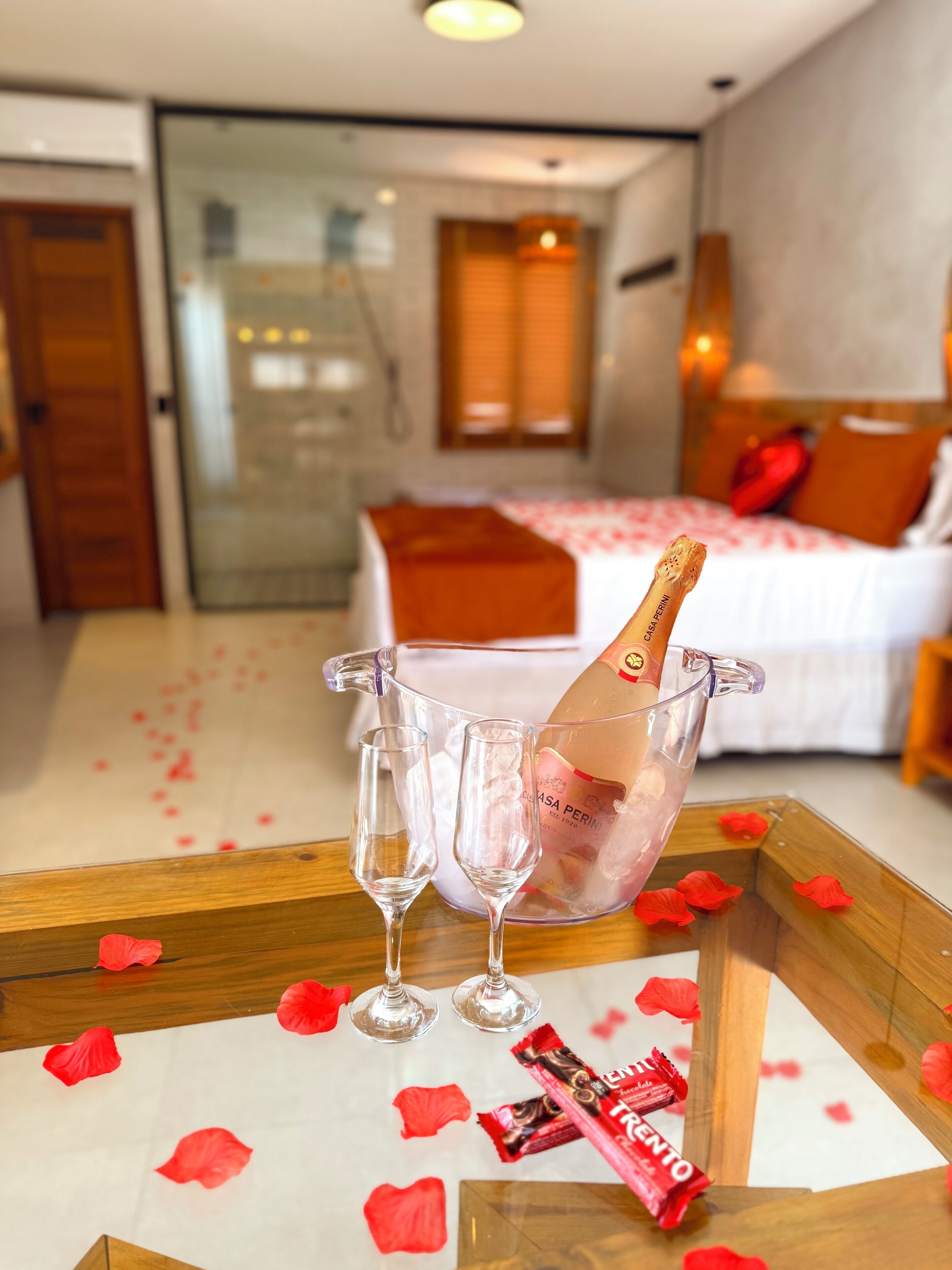 Una habitación con una cama, una mesita de noche con una botella de champán, dos copas y pétalos de rosa en el suelo.