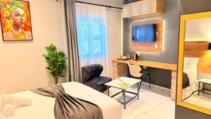 Quarto de hotel com cama, escrivaninha, cadeira, televisão e espelho.