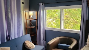 Una habitación con un sofá azul, un sillón de cuero, una cama, un ventanal con vistas a la vegetación y un cuadro en la pared.
