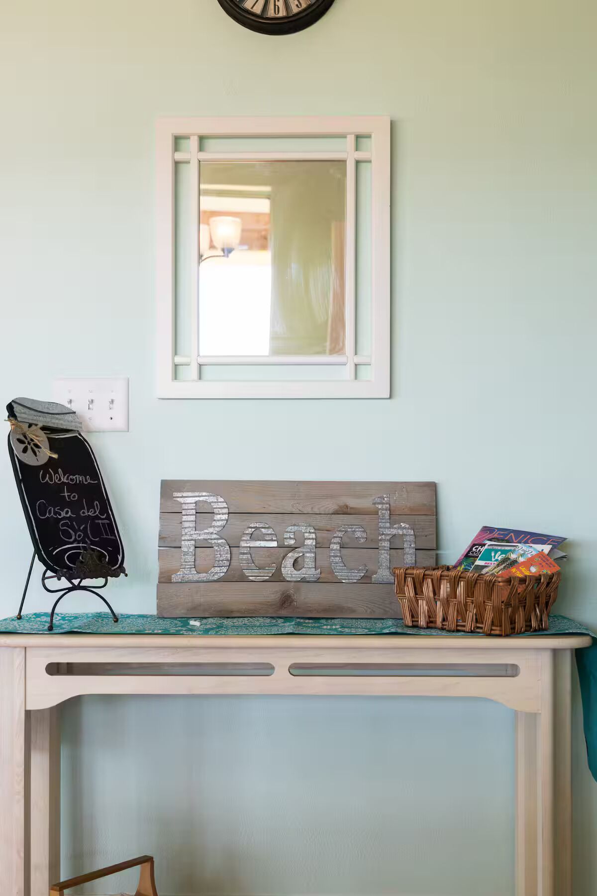Une horloge murale, un miroir encadré, un panneau noir, une enseigne en bois et un panier en osier contenant des magazines, le tout posé sur une petite table console de couleur claire.
