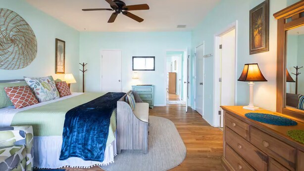 Un dormitorio con una cama, mesitas de noche, un aparador, un ventilador de techo y una puerta que da a otra habitación.