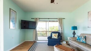 Un salón con un sillón azul, un sofá beige, un televisor de pantalla plana montado en la pared y un ventanal con cortinas.
