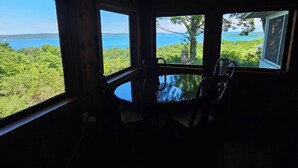 Een kamer met een ronde tafel en stoelen, grote ramen met uitzicht op een bos en een waterpartij.