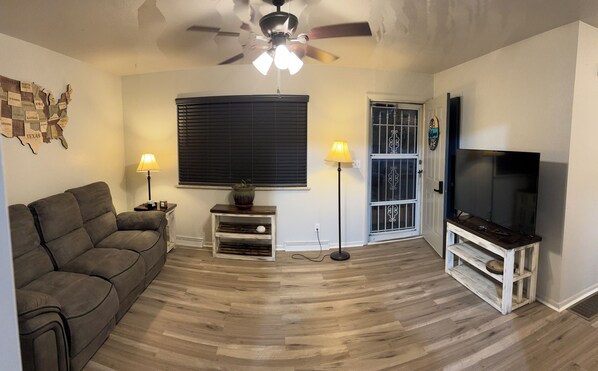 A living room with a sofa, a television, two lamps, a ceiling fan, and a window with blinds.
