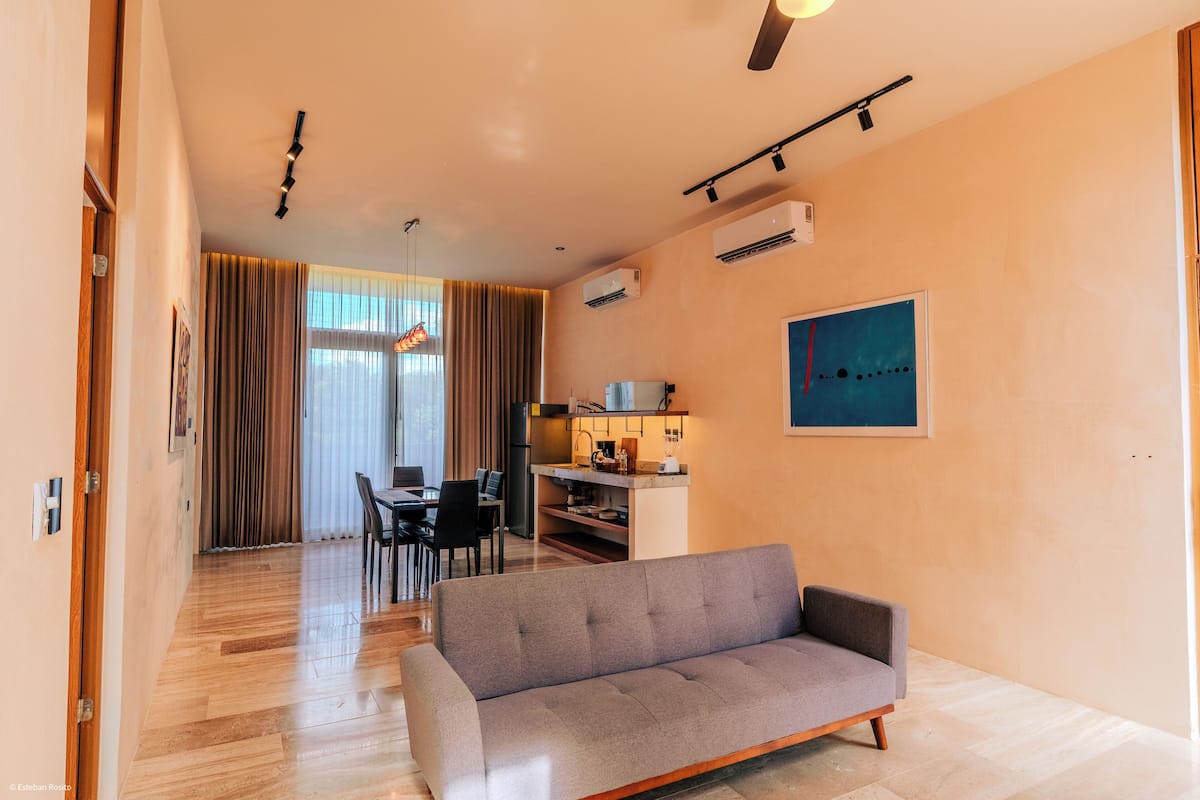A modern living room with a grey sofa, wooden flooring, a dining area with chairs, a kitchen with a countertop, and a ceiling fan.
