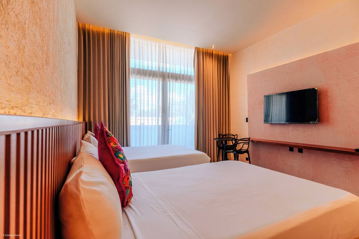 A hotel room with two beds, a TV mounted on the wall, and a window with curtains.