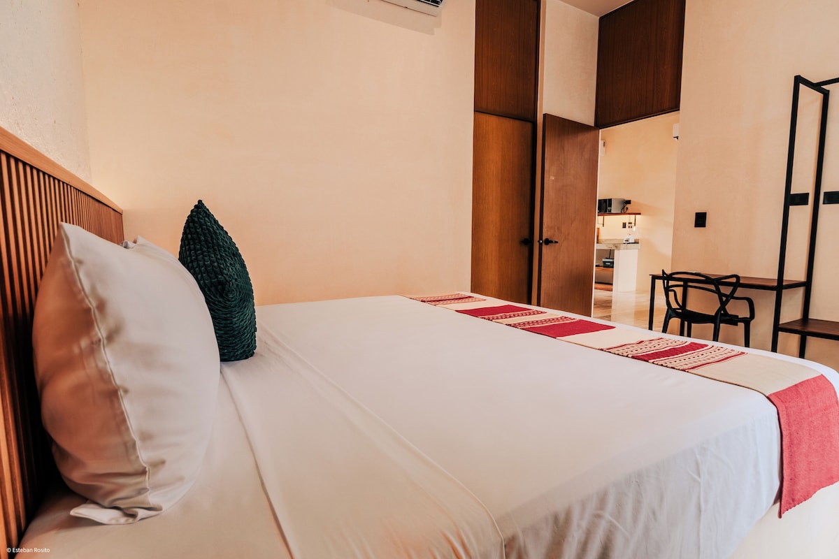 A neatly made bed with a white bedspread and red decorative pillows, a wooden headboard, and a small table with chairs in the background.
