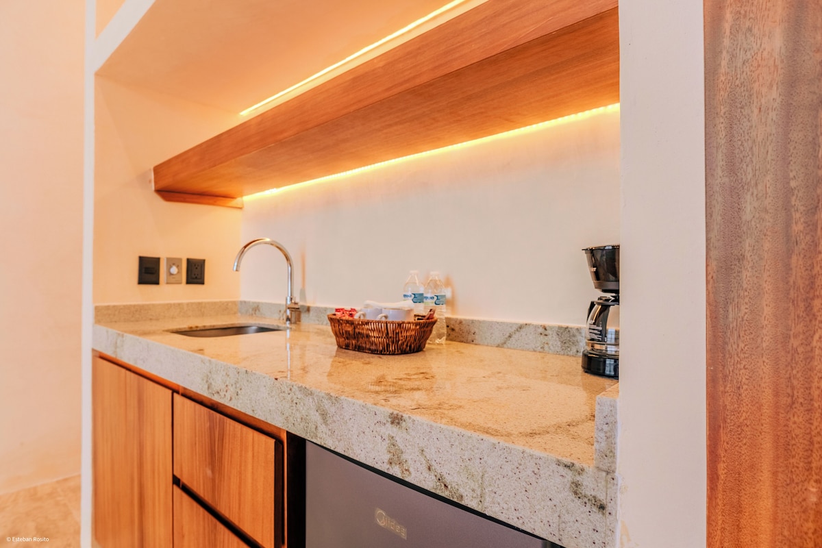A kitchen with a granite countertop, a stainless steel sink, a coffee maker, and a basket with bottles.