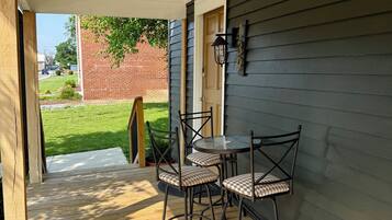 A covered patio with a table and chairs, a wooden deck, and a view of a brick building and greenery.