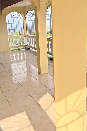 A spacious indoor area with tiled flooring, a balcony with a white balustrade, and large windows with metal bars. - Moorish Mount King Sanctuary – Hilltop Views, Private Bath, Mandeville (Mandeville)