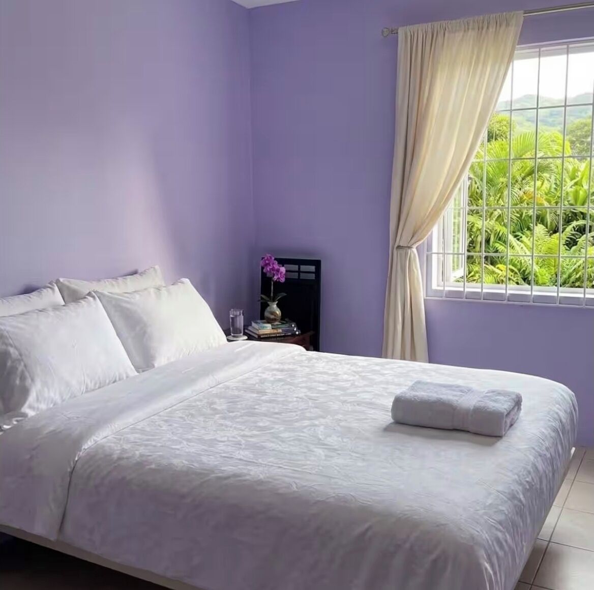 A bedroom with a bed, a nightstand, a window with curtains, and a view of greenery outside.