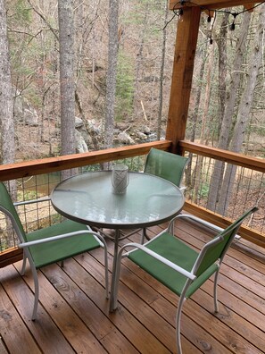 Eine Holzterrasse mit einem Glastisch und vier grünen Stühlen, mit Blick auf eine bewaldete Gegend.