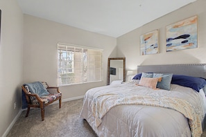 Une chambre avec un grand lit, une chaise, une fenêtre avec des stores et deux tableaux accrochés au mur.