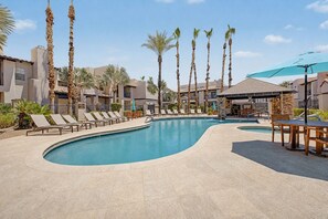 A swimming pool with lounge chairs and palm trees, surrounded by buildings with a Mediterranean architectural style.