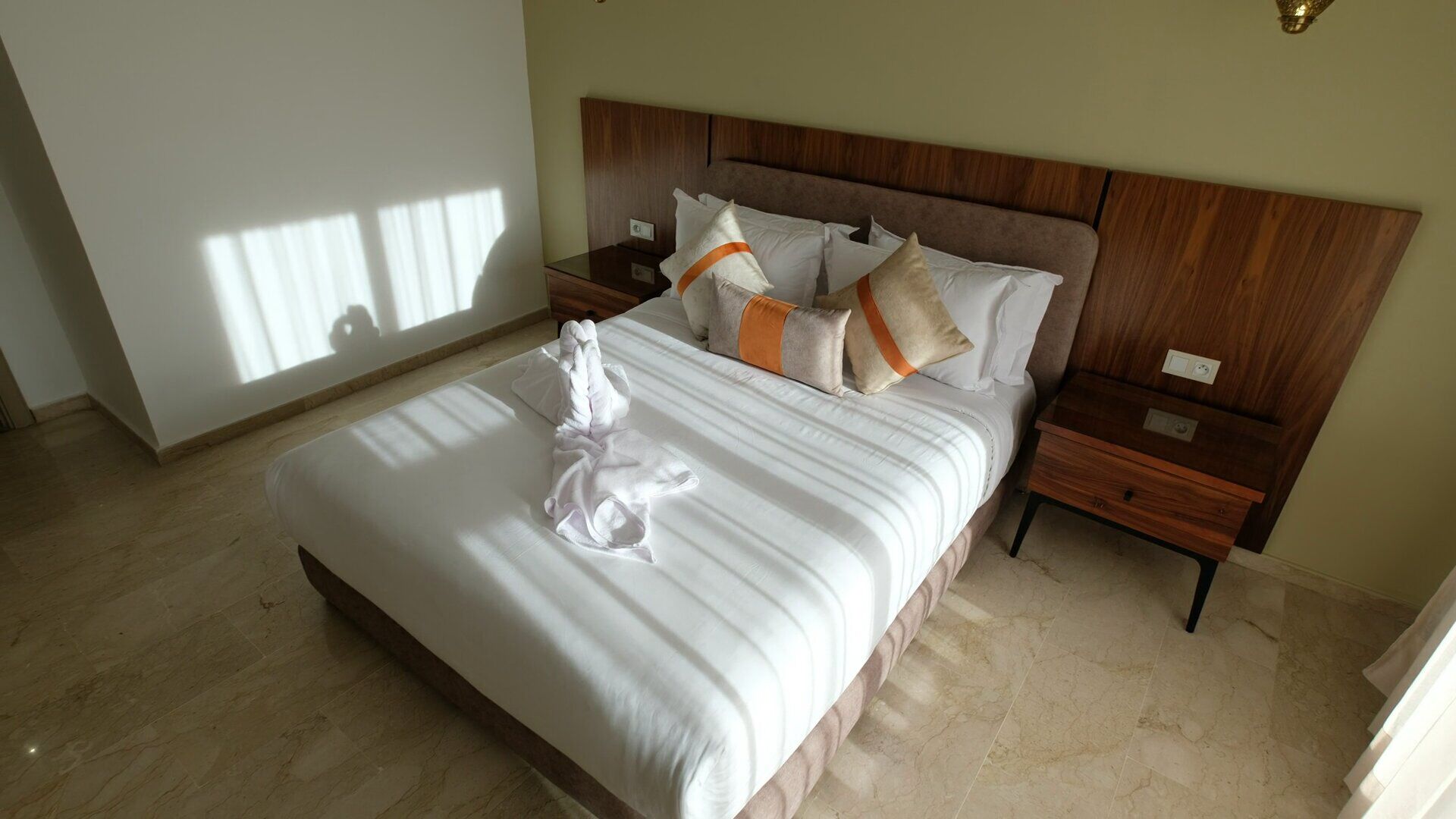 A neatly made bed with white linens and a folded towel, a wooden headboard, and a nightstand with a lamp.