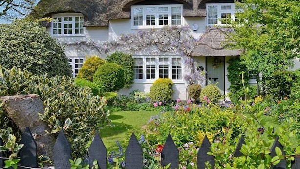 Una casa tradicional con techo de paja, paredes blancas y múltiples ventanas, rodeada por un jardín cuidado con diversas plantas y una valla de piquetes negros.