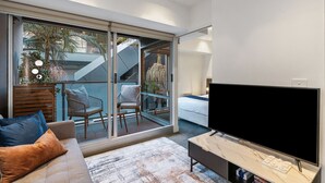 A modern living room with a large flat-screen TV, a sofa with cushions, a coffee table, and a sliding glass door leading to an outdoor balcony with chairs and plants. - The Inkerman Hideaway (St Kilda)