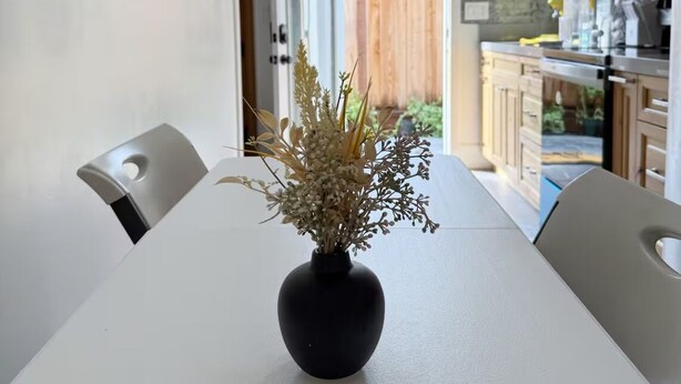 A dining table with a black vase of dried flowers in the center, white walls, and wooden cabinets in the background.