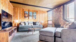 A cozy living room with a wooden paneled wall, a stone accent wall, a large gray sofa, a coffee table, and a TV on a wooden stand.