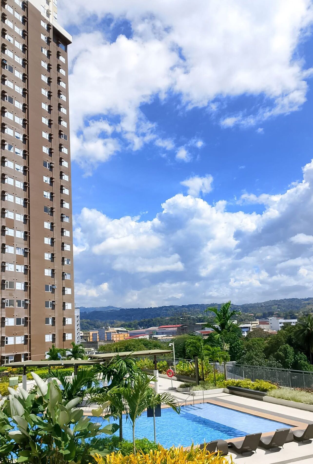 A high-rise building with a pool area, surrounded by greenery and a clear sky.