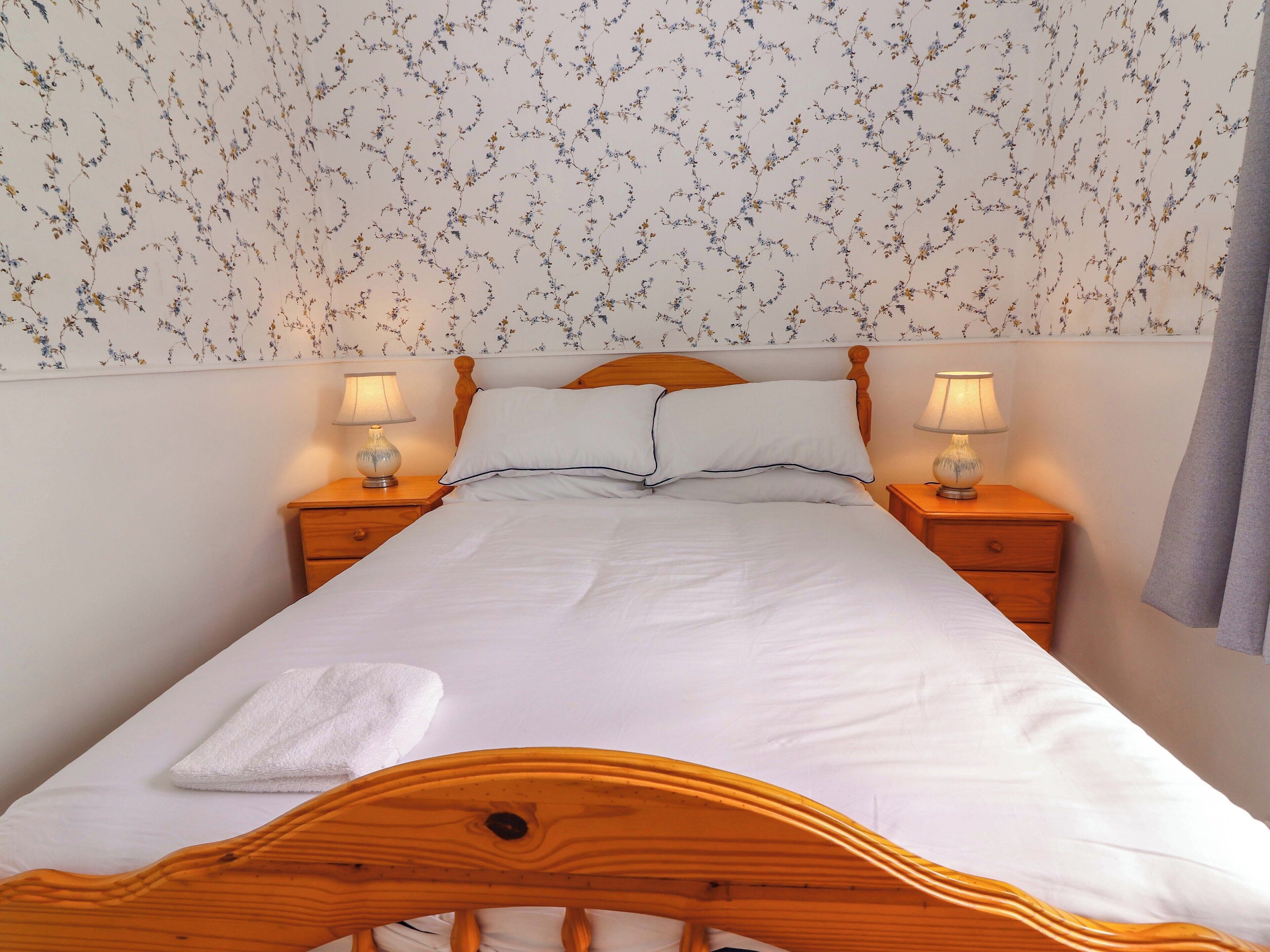 A neatly made bed with white linens and pillows, flanked by two bedside tables with lamps, in a room with floral wallpaper.