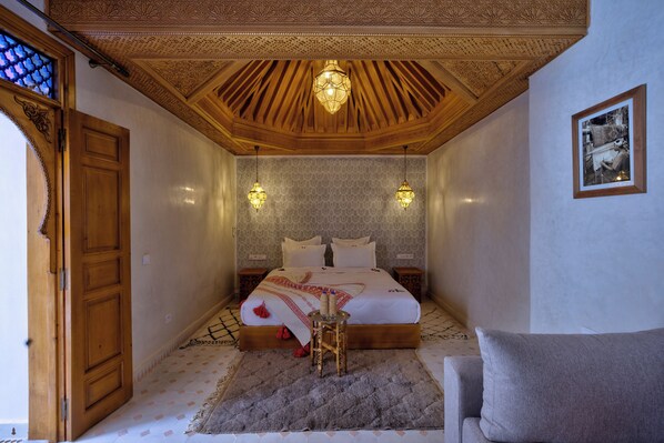 A bedroom with a wooden ceiling, a bed with a patterned bedspread, two wall-mounted lamps, and a framed picture on the wall.