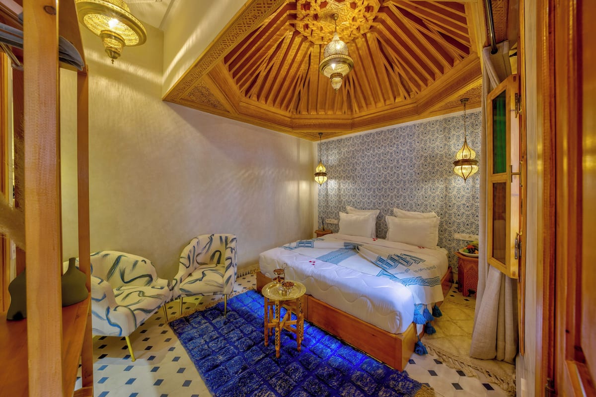 A bedroom with a large bed, a chair, a small table, and a decorative ceiling.