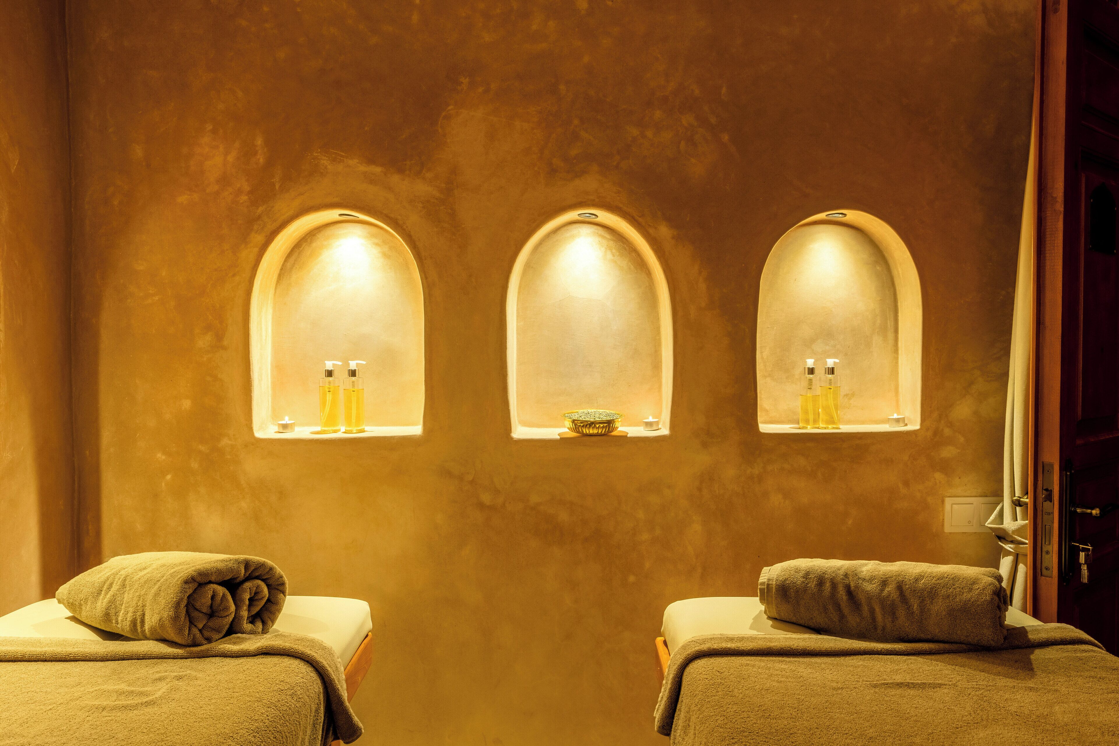 Un cuarto de spa con tres nichos arqueados que contienen botellas, un par de toallas sobre mesas de masaje, y una puerta de madera.