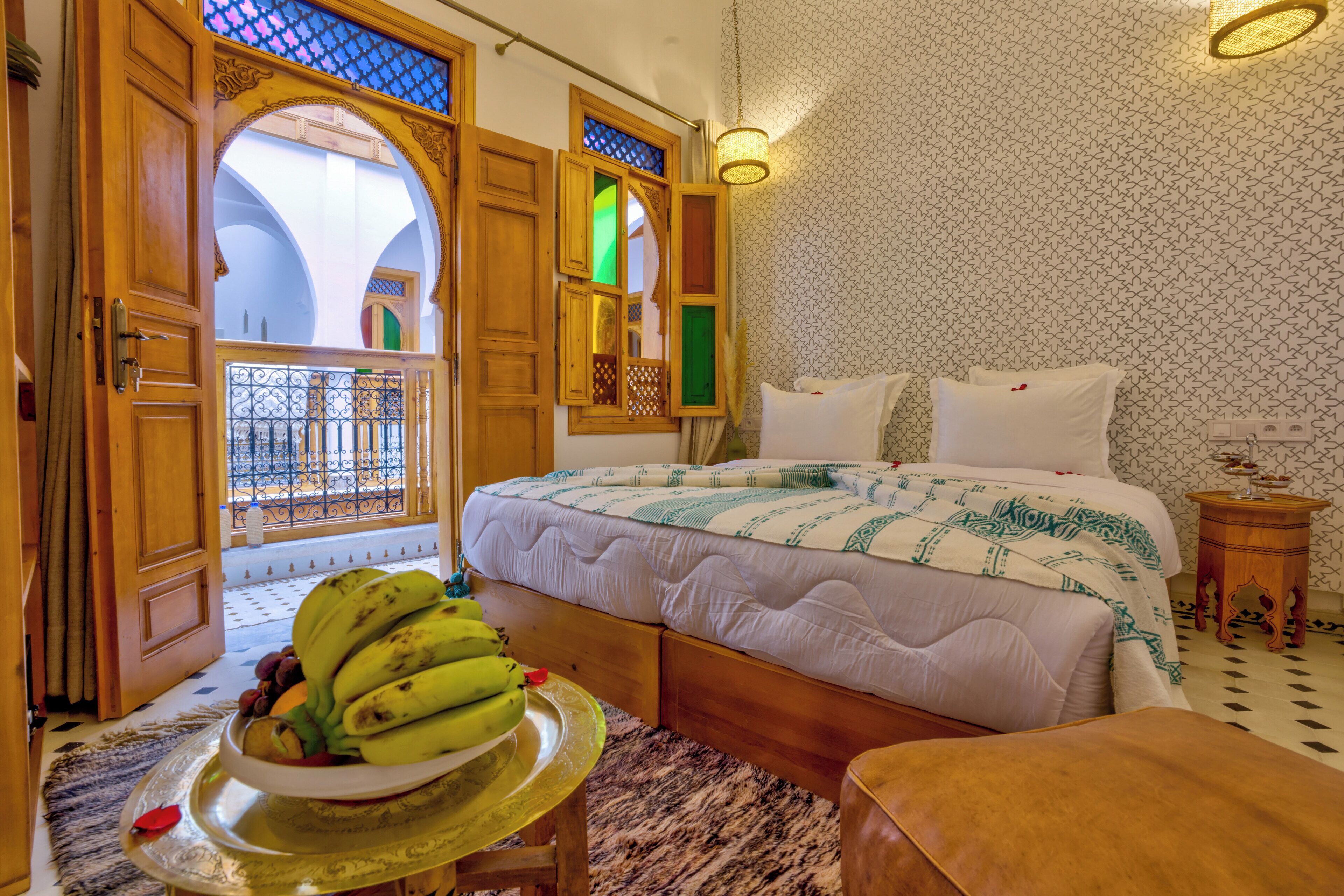 A bedroom with a large bed, a wooden door, a balcony with a view, and a fruit bowl on a table.