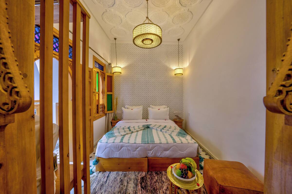 A bedroom with a bed, wooden headboard, patterned wallpaper, and a hanging light fixture.