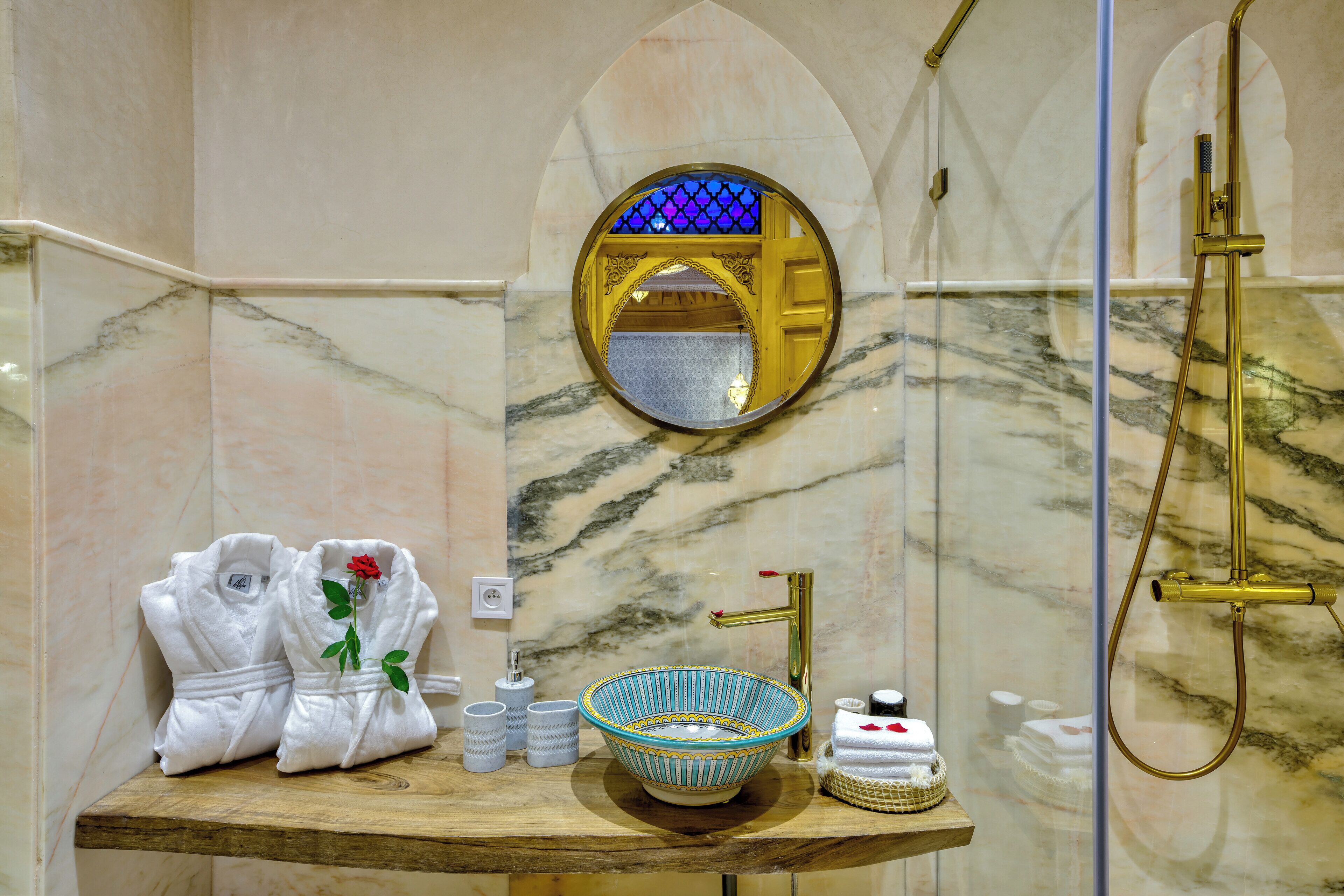 Une salle de bain avec un lavabo en marbre, un robinet doré, un miroir rond et deux peignoirs blancs ornés d’une rose.