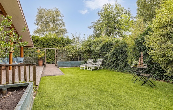 A backyard with a wooden deck, two lounge chairs, a small table, and a garden area with various plants and trees.