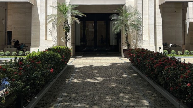 Entrada de un edificio con el letrero 'WAVE IPANEMA' sobre las puertas, flanqueada por palmeras y macizos de flores.