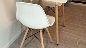 Un coin repas avec une table et des chaises blanches, un plancher en bois et une vue sur l’extérieur par une fenêtre agrémentée de rideaux.