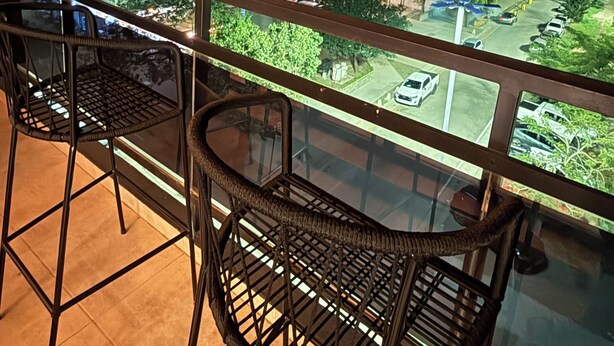 Un balcon avec deux fauteuils en osier, une vue sur le paysage urbain illuminé la nuit, et un immeuble dont les fenêtres sont éclairées.