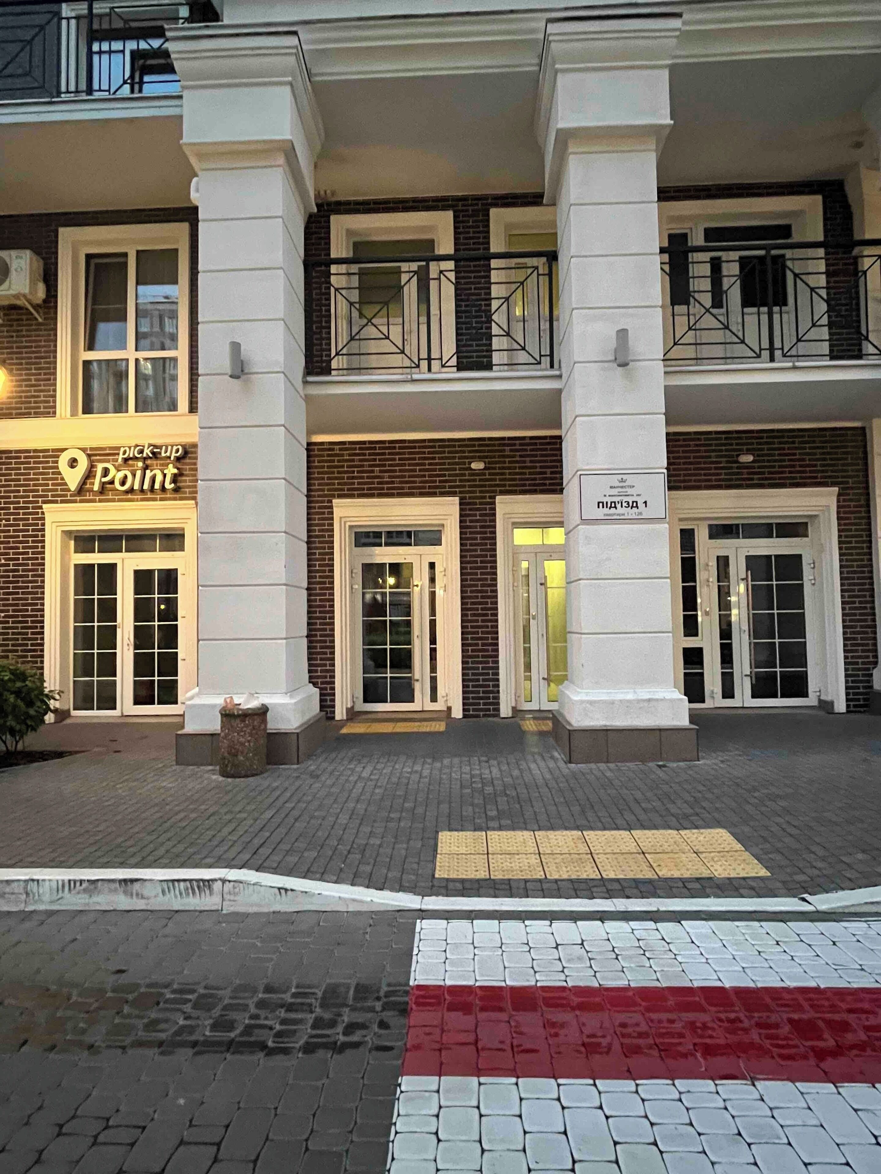 Um edifício com placa indicando 'ponto de recolha', portas duplas e uma rampa com piso tátil.