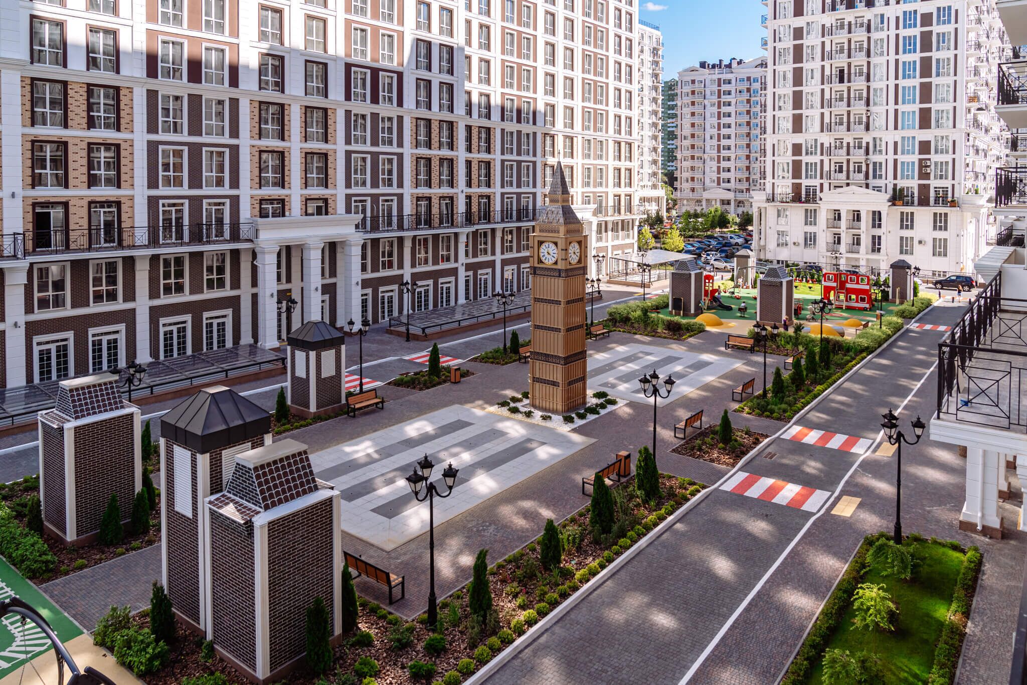 A model of a residential building complex with a central clock tower, multiple balconies, and a pedestrian area with benches and street lamps.