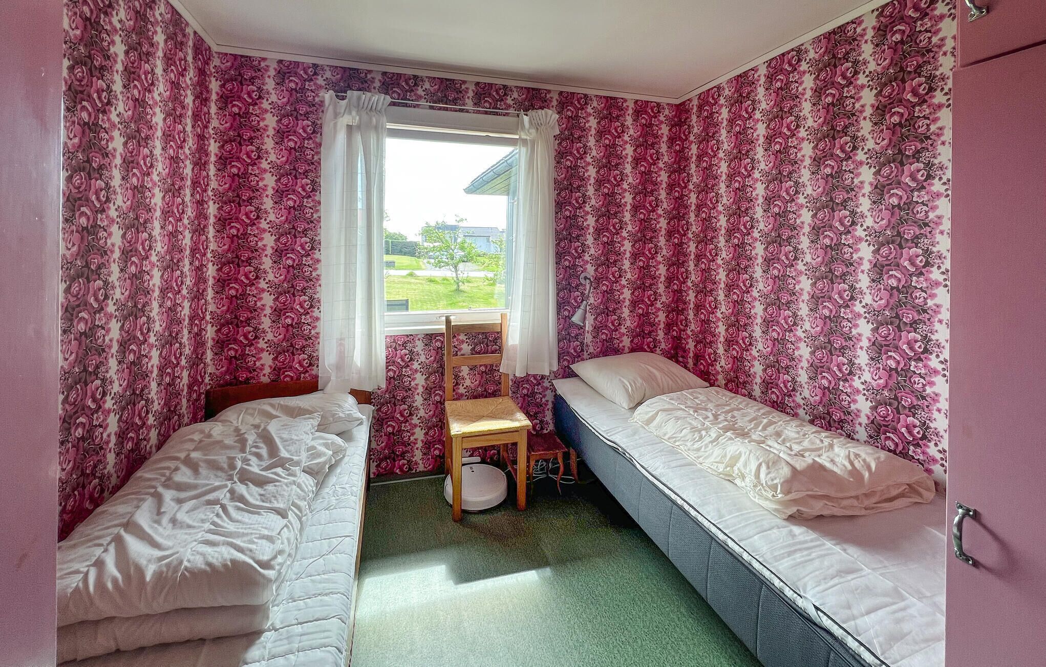 Ett sovrum med två enkelsängar, en liten trästol, ett fönster med vita gardiner och utsikt över grönområden utanför.
